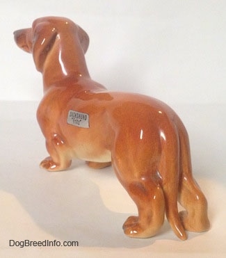 The back left side of a brown Dachshund figurine that is in a standing pose. The figurines ears are attached to its neck.