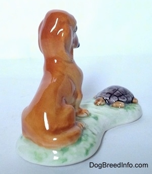 The back right side of a figurine of a brown Dachshund that is sitting across and looking down at a tortoise in front of it. The Tortoise figurine is looking at the Dachshund in front of it.