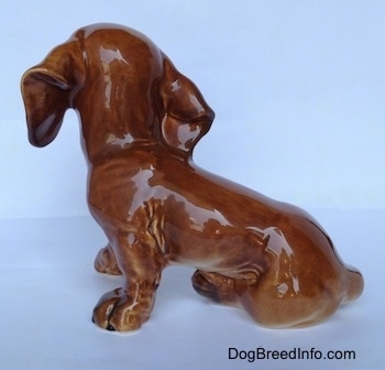 The left side of a brown Dachshund puppy figurine that is in a sitting pose. The figurine has short legs and it is glossy.