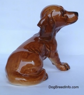 The back right side of a figurine of a brown Dachshund puppy. The figurine has a long tail that wraps around its leg.