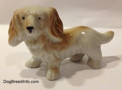 The left side of a white with tan porcelain long haired Dachshund figurine. The figurine has long hair details and it has tiny black circles for eyes.