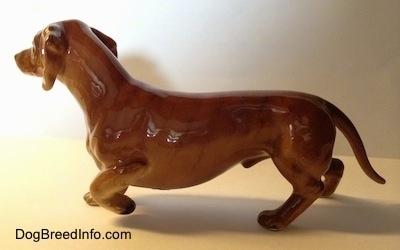 The left side of a brown porcelain Dachshund pointing figurine. The figurine has a long body, short legs and a long tail.