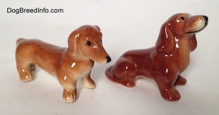 The front right side of two diifferent Dachshund figurines. Both Dachshunds have black circles for eyes.