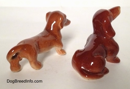 The back right side of a sitting Dachshund and a standing Dachshund figurine. The figurines are glossy.