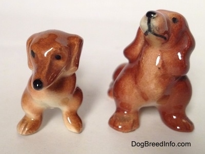 Two different Dachshund figurines, one figurine is sitting and the other is standing. The figurines have short legs.