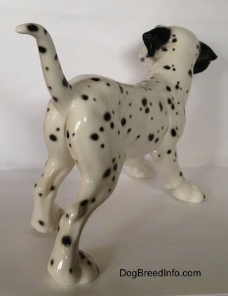 The back right side of a Dalmatian figurine. The figurine is glossy and it has black ears. The tail is long and up in the air. The dog has black spots all over its body.