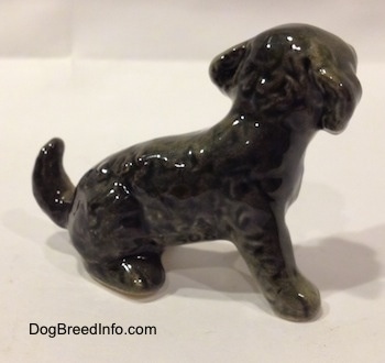 The right side of a figurine that is a black and gray Dandie Dinmont Terrier puppy in a sitting position. The figurine has a short arching up tail.