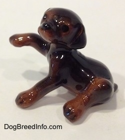 The left side of a figurine that is a miniature black and brown Doberman Pinscher puppy with its paw up. The figurine has legs as long as its body.