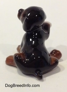 The back side of a figurine that is a miniature black and brown Doberman Pinscher puppy with its paw up. The figurine has a short tail.