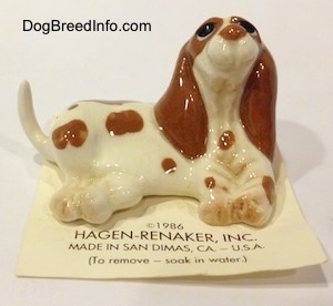 A figurine of a white with brown Curbstone Setter in a lying pose. The figurine is laying on top of a card and its head is in the air.
