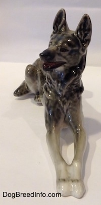 A porcelain black with gray and white East-European Shepherd lying figurine. The figurines head is turned to the left and its mouth is open.