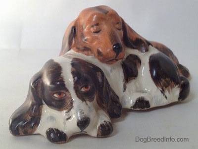 The front left side of a figurine that is two English Cocker Spaniel dogs that are laying down. The figurines are different colors the dog on the bottom is brown and white and the dog on top is orange and black.