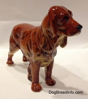 The front right side of a red figurine of a English Cocker Spaniel. The figurine has long legs and small paws.