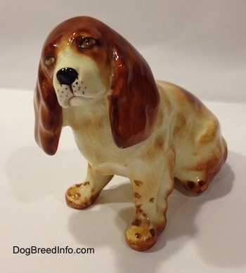 The front left side of a red and white ceramic English Cocker Spaniel figurine in a sitting position. The figurine has large big ears.