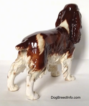 The back right side of a brown and white English Springer Spaniel in a standing pose figurine. The figurine has a long body, long legs and small brown paws.