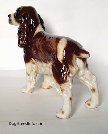 The back left side of a figurine of a brown and white English Springer Spaniel figurine in a standing pose. The figurine has a small brown spots going down its legs.