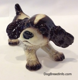 A figurine of a liver and white English Springer Spaniel puppy with its ears flying out. The figurine has black circles for eyes.