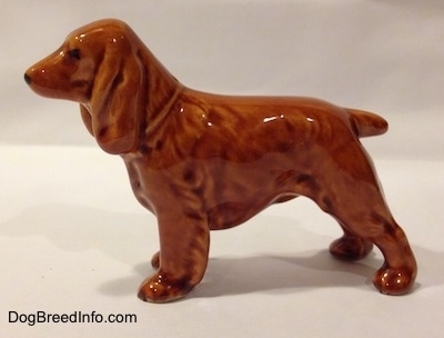 The left side of a brown ceramic Field Spaniel figurine dog. The figurine has large brown ears a short docked tail and a pointy muzzle.