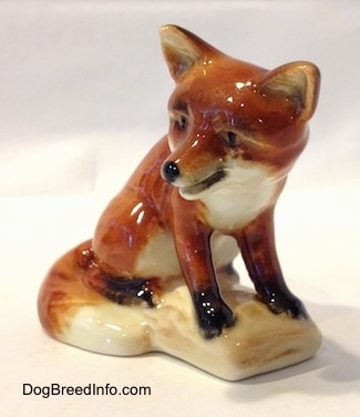 The front right side of a porcelain figurine of a red fox sitting on a log. The figurine has black paws. Its mouth is open.
