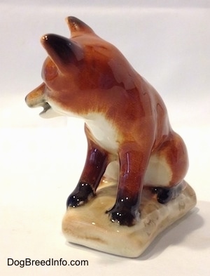 The front left side of a red fox porcelain figurine that is sitting on a log. The figurine has a white chest and the back of its ears are black.