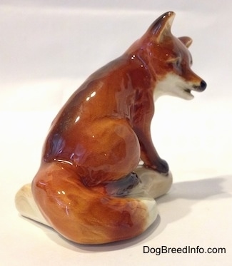The back right side of a porcelain figurine of a red fox. The figurine has its mouth slightly open and its tail is around a log.