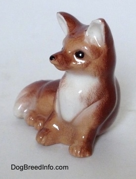 The front left side of a baby Fox seated figurine. The Fox has a white chest and a white tipped tail.