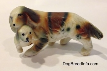 The left side of a brown and white with black bone china of a French Brittany Spaniel carrying a puppy. Both parts of the figurine have black circles for eyes.