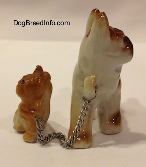 A brown with white and black French Bulldog has a chain that connects it to a brown with tan Frenchie puppy figurine. The figurines both have brown paws.