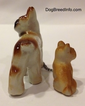 The back of a brown with white and black French Bulldog figurine has a chain on it that connects it to a brown with tan Frenchie puppy figurine that is sitting. Both figurines have short tails.