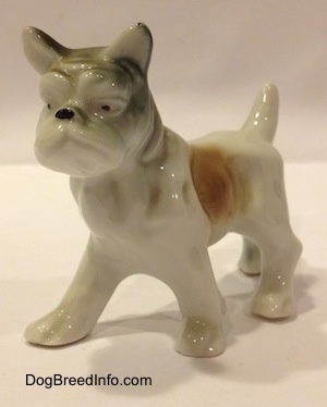 The front left side of a white with brown and green porcelain French Bulldog figurine in a standing pose. The figurine has mediumg sized legs and small paws.