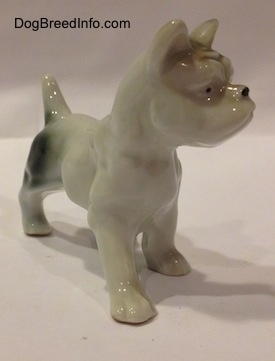 The front right side of a porcelain figurine of a white with brown and green French Bulldog in a standing pose. The figurine has small black circles for eyes.