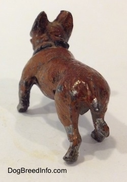 The back left side of a figurine of a brown with black French Bulldog that is made of metal. The figurine has a short tail with chipped paint towards the end.