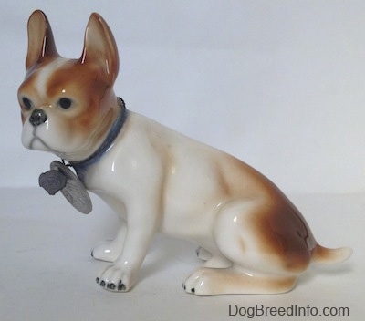 The left side of a brown and white French Bulldog figurine in a sitting pose. The figurine is wearing a blue collar and it has a large medallion on the collar.