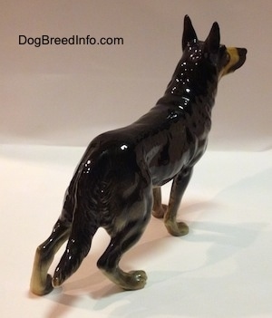 The back right side of a black with tan standing German Shepherd figurine. The figurine has a long fluffy tail.