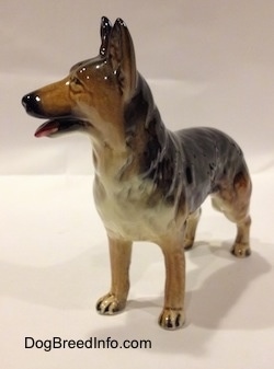 The front left side of a figurine of a porcelain black with brown and white German Shepherd standing. The figurine has its mouth open.
