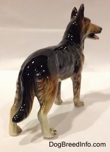 The back right side of a figurine of a porcelain black with brown and white German Shepherd standign. The figurine has a long tail.
