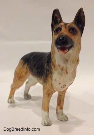 A porcelain figurine of a black and tan with white standing German Shepherd. The figurines mouth is open and tongue is sticking out.