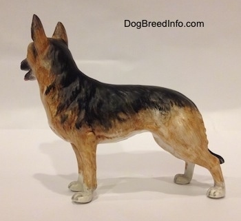 The left side of a porcelain black and tan with white figurine of a German Shepherd standing. The figurine has fine hair details along its body.