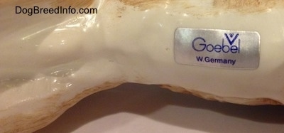 The underside of a realistic porcelain German Shepherd figurine and on the underside is a sticker with the logo of Goebel W.Germany on it.