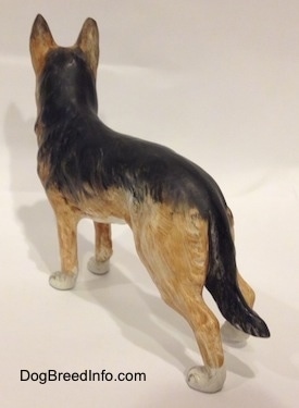 The back left side of a porcelain figurine of a black and tan with white standing German Shepherd. The figurine has its ears in the air.