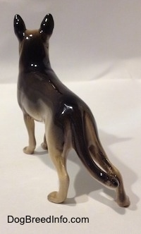 The back left side of a figurine of a black with tan German Shepherd standing. The figurine has long legs and the back right one sticks out.