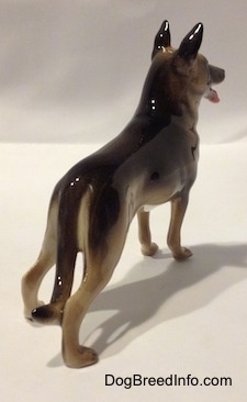 The back right side of a black with tan figurine of a German Shepherd. The figurines tail is long.