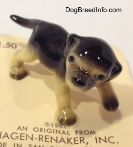 The front left side of a black with tan figurine of a German Shepherd puppy. The figurine has its head tilted and big black circles for eyes.