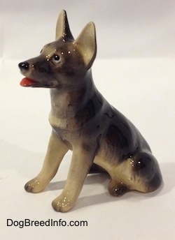 The front left side of a black with tan sitting German Shepherd figurine. The figurine has black circles for eyes with a small white circle inside of it.