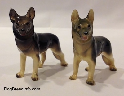 The front left side of two black with tan German Shepherd standing figurines. The figurines have black circles for eyes and a nose.