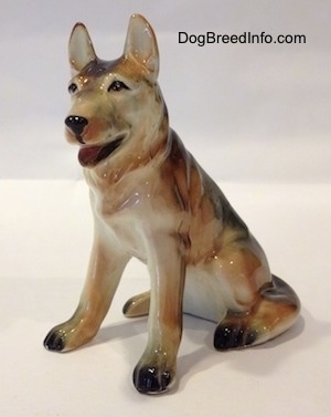 The front left side of a ceramic figurine of a black with tan and white German Shepherd sitting. The figurines ears are standing.