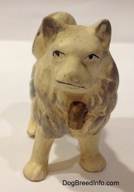 A white with gray German Spitz ceramic figurine. The figurine has black circles for eyes and it has an item on its chest.