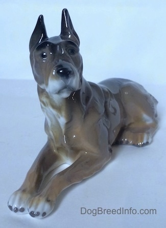The front left side of a Great Dane figurine in a laying down pose. The figurine has black circles for eyes and its ears are sticking up.