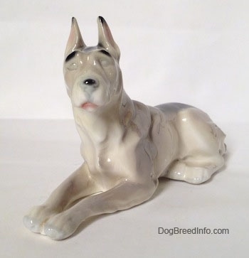 The front left side of a figurine of a white with black Great Dane laying down figurine. The figurine has long legs and small paws.