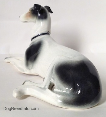 The back left side of a white with black and tan Greyhound figurine in a lying pose. The figurine has a large tail.